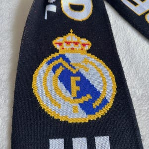 May include: Black scarf with the Real Madrid logo, featuring a gold crown and emblem with the letters "RM" in blue. The scarf has white text on a black background.