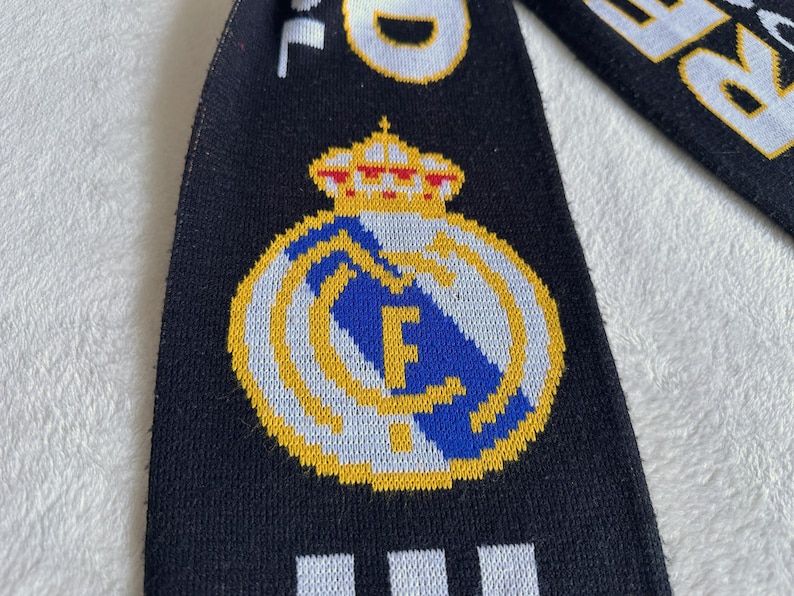 May include: Black scarf with the Real Madrid logo, featuring a gold crown and emblem with the letters "RM" in blue. The scarf has white text on a black background.