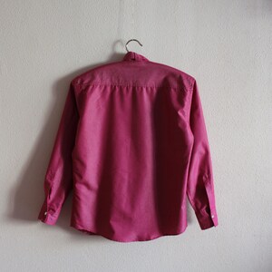 May include: A long-sleeved, button-down shirt in a deep pink color. The shirt is hanging on a hanger against a white wall.