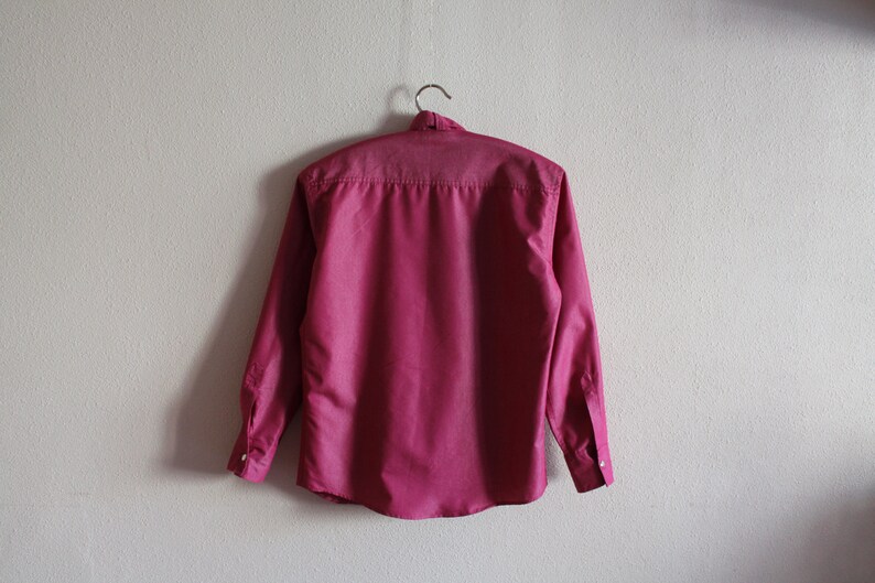May include: A long-sleeved, button-down shirt in a deep pink color. The shirt is hanging on a hanger against a white wall.