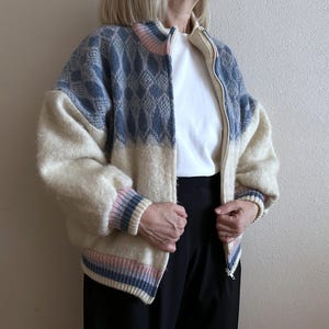 May include: A zip-up jacket with a cream-colored body and blue patterned shoulders. The jacket has a high collar and ribbed cuffs with pink and blue stripes. The model is wearing a white t-shirt and black pants.