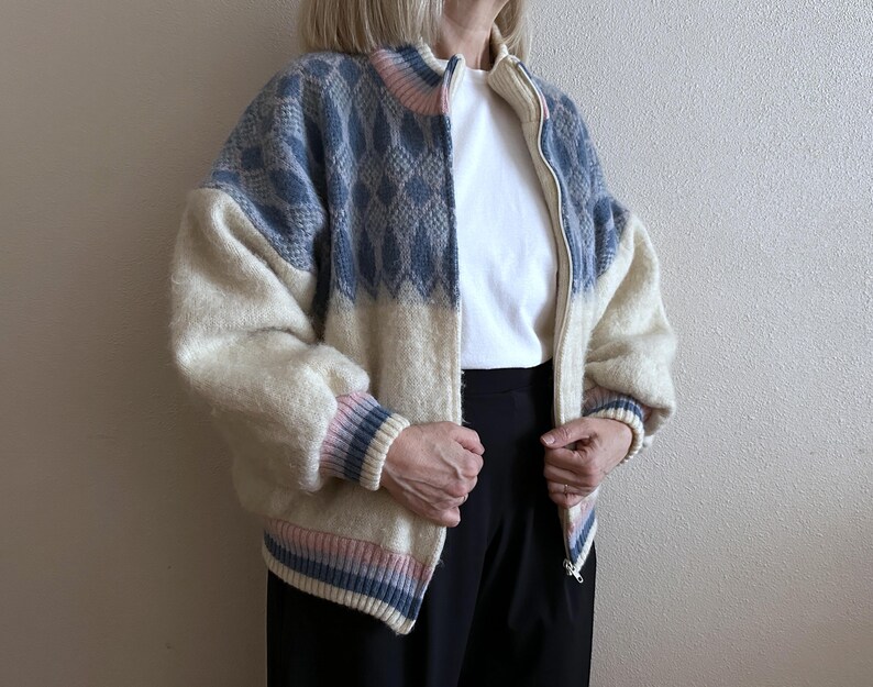 May include: A zip-up jacket with a cream-colored body and blue patterned shoulders. The jacket has a high collar and ribbed cuffs with pink and blue stripes. The model is wearing a white t-shirt and black pants.