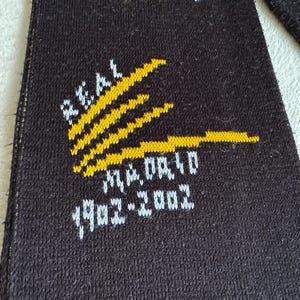 May include: Black fabric with the words "REAL MADRID" in white and yellow. The text "1902-2002" is also visible. The design features yellow stripes.