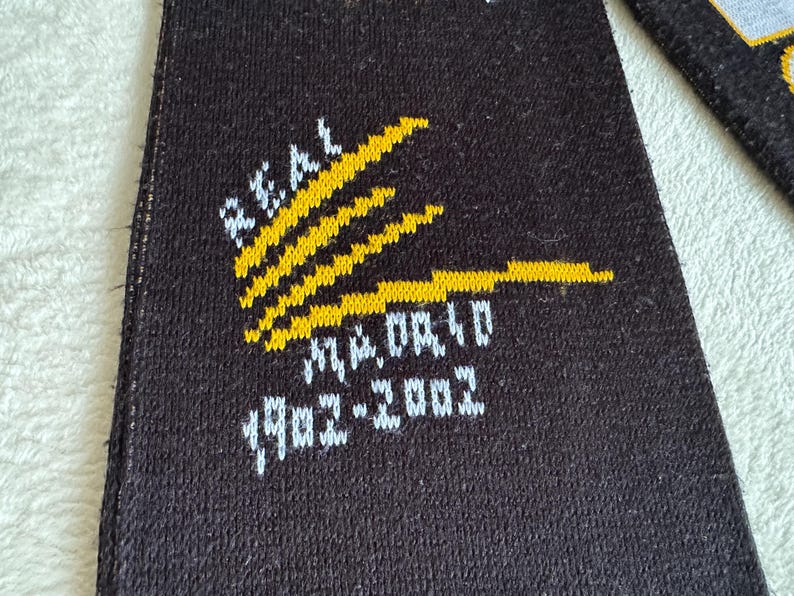 May include: Black fabric with the words "REAL MADRID" in white and yellow. The text "1902-2002" is also visible. The design features yellow stripes.