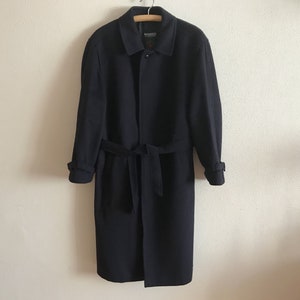 Könnte beinhalten: Ein marineblauer Mantel mit passendem Gürtel, der an einem Holzbügel hängt. Der Mantel hat ein klassisches Design mit Kragen, Knopfverschluss und langen Ärmeln. Der Markenname "Bond & Co" ist auf dem Etikett zu sehen.