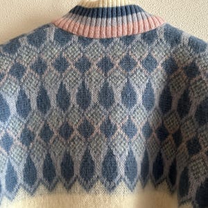 May include: Close-up of a cozy sweater with a diamond pattern. The sweater features a cream-colored base with a repeating pattern of blue and light pink diamonds. The collar is ribbed with blue and pink stripes.