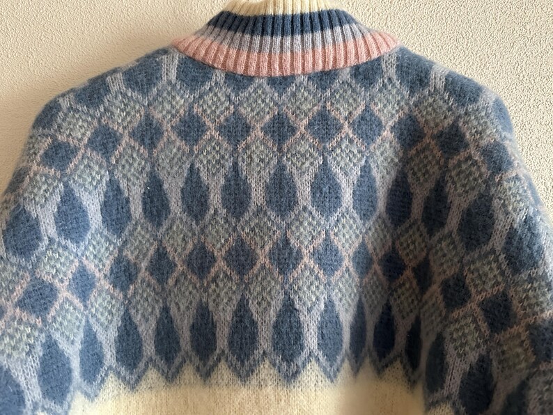May include: Close-up of a cozy sweater with a diamond pattern. The sweater features a cream-colored base with a repeating pattern of blue and light pink diamonds. The collar is ribbed with blue and pink stripes.