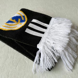 May include: Folded black scarf with white fringe. The scarf features a team logo in blue, yellow, and white, along with white stripes. The scarf is made of a knitted material.