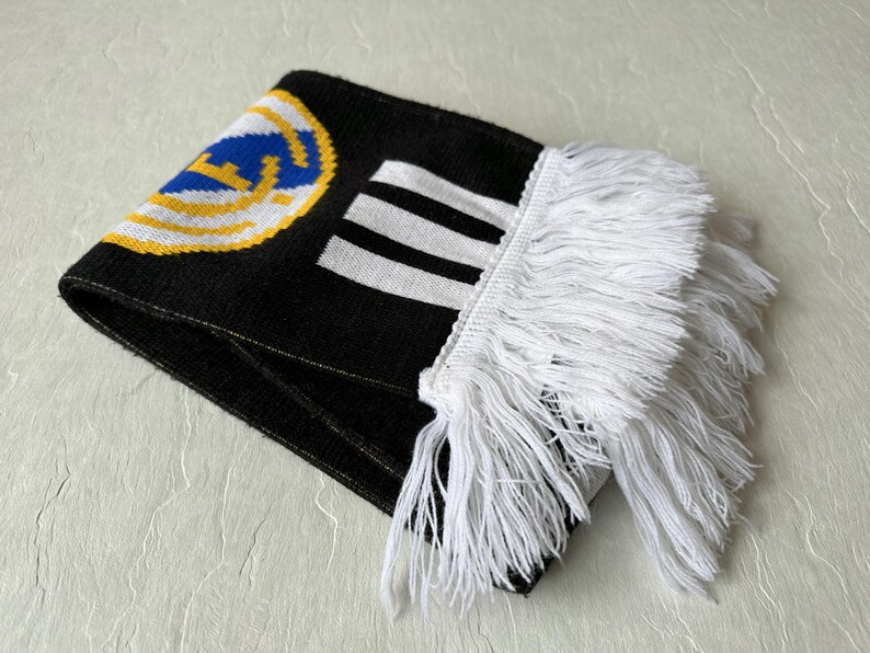 May include: Folded black scarf with white fringe. The scarf features a team logo in blue, yellow, and white, along with white stripes. The scarf is made of a knitted material.