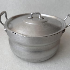 Puede incluir: Una olla de cocina de color plateado con tapa. La olla tiene dos asas curvas a los lados y un asa pequeña en la tapa. La olla parece ser de metal y está diseñada para cocinar.