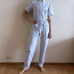 Könnte beinhalten: Hellblauer Overall mit kurzen Ärmeln, Reißverschluss vorne und passendem Gürtel. Der Overall hat einen kleinen gestickten Aufnäher auf der Brusttasche. Das Outfit ist aus einem hellen Stoff gefertigt.