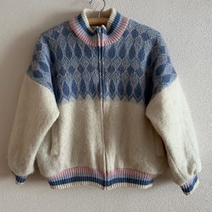 May include: A vintage zip-up jacket with a blue and white diamond pattern on the upper chest and sleeves. The jacket has a cream-colored body, a ribbed collar, cuffs, and hem with pink and blue stripes. The jacket is hanging on a wooden hanger.