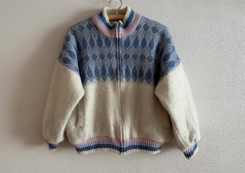 May include: A vintage zip-up jacket with a blue and white diamond pattern on the upper chest and sleeves. The jacket has a cream-colored body, a ribbed collar, cuffs, and hem with pink and blue stripes. The jacket is hanging on a wooden hanger.