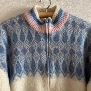 May include: A zip-up sweater with a high collar. The sweater features a blue and white diamond pattern on the upper portion and white on the lower portion. The collar is striped with white, pink, and blue. The sweater is hanging on a wooden hanger.