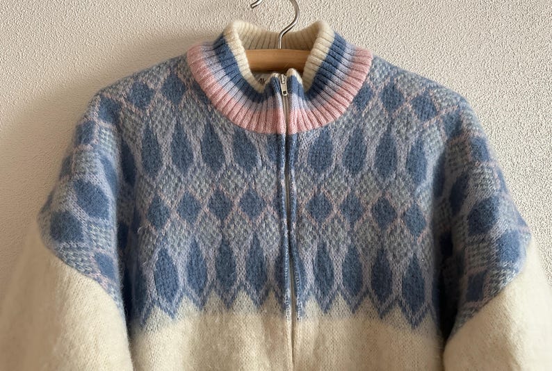 May include: A zip-up sweater with a high collar. The sweater features a blue and white diamond pattern on the upper portion and white on the lower portion. The collar is striped with white, pink, and blue. The sweater is hanging on a wooden hanger.