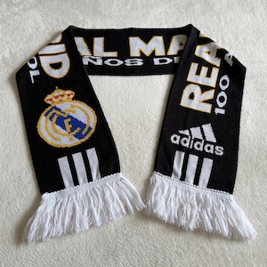 May include: Black and white knitted scarf with white fringe. The scarf features the Real Madrid crest, the Adidas logo, and text in white and gold. The scarf is a fan accessory.