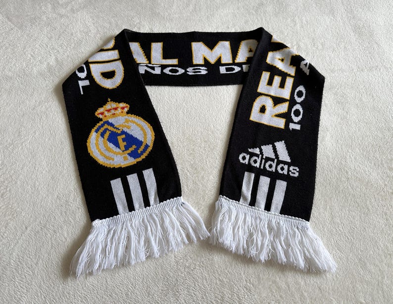 May include: Black and white knitted scarf with white fringe. The scarf features the Real Madrid crest, the Adidas logo, and text in white and gold. The scarf is a fan accessory.