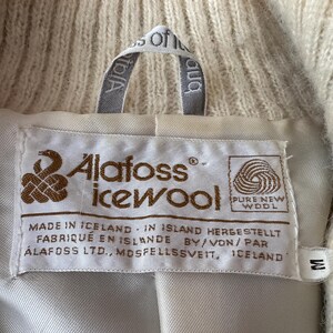 May include: Close-up of a cream-colored wool garment label. The label reads "Alafoss icewool" with text in English, German, and French. The garment is made in Iceland and is size M.