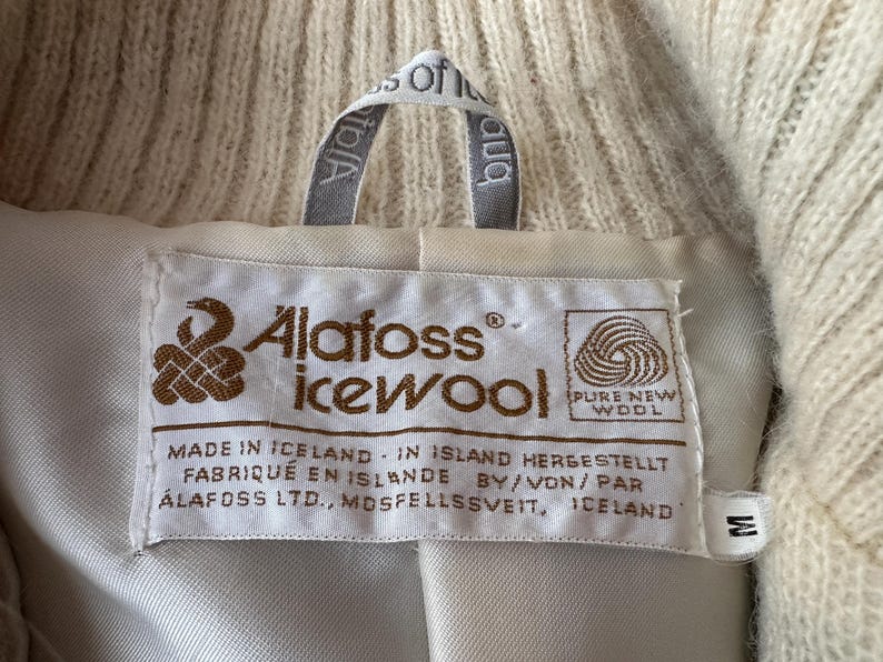 May include: Close-up of a cream-colored wool garment label. The label reads "Alafoss icewool" with text in English, German, and French. The garment is made in Iceland and is size M.
