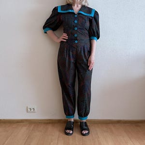 Może przedstawiać: Vintage kombinezon z wzorem paisley w odcieniach czerni, zieleni i czerwieni. Kombinezon ma duży kołnierz i mankiety wykończone turkusowym niebieskim, z pasującymi guzikami. Ubranie ma dopasowany krój i jest noszone z czarnymi sandałami. Wymiary w centymetrach.
