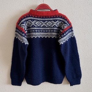 Puede incluir: Un suéter azul marino con cuello y hombros rojos. El suéter presenta un patrón geométrico blanco en el pecho y los hombros. Cuelga de una percha roja.