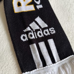 May include: Black and white Adidas scarf with the Adidas logo and three white stripes. The scarf has white fringe at the bottom and the text "R20" in white and yellow.