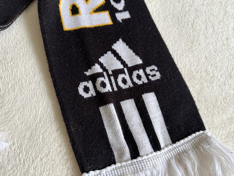 May include: Black and white Adidas scarf with the Adidas logo and three white stripes. The scarf has white fringe at the bottom and the text "R20" in white and yellow.