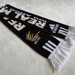 May include: Black scarf with white and gold text, featuring the word "REAL" and the Adidas logo. The scarf has white fringe at the ends and text that reads "100 A&Ntilde;OS" and "REAL MADRID 1902-2002".