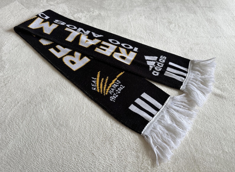 May include: Black scarf with white and gold text, featuring the word "REAL" and the Adidas logo. The scarf has white fringe at the ends and text that reads "100 A&Ntilde;OS" and "REAL MADRID 1902-2002".