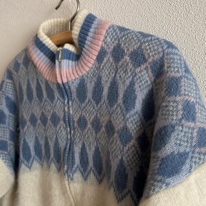 May include: A vintage zip-up sweater with a diamond pattern in shades of blue and gray. The collar features a gradient of pink and blue. The lower portion and sleeves are off-white. The sweater is hanging on a wooden hanger.