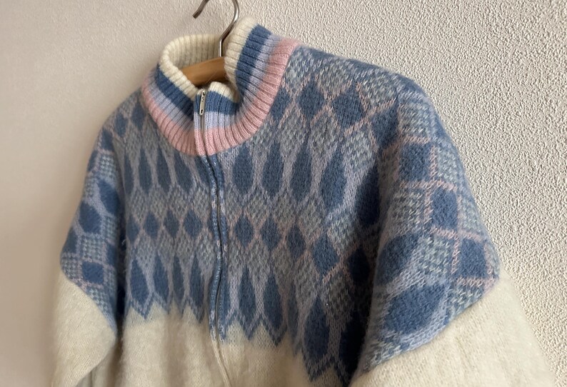 May include: A vintage zip-up sweater with a diamond pattern in shades of blue and gray. The collar features a gradient of pink and blue. The lower portion and sleeves are off-white. The sweater is hanging on a wooden hanger.