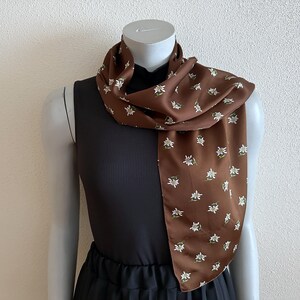 May include: A brown scarf with a white floral pattern is draped around a mannequin's neck. The scarf is made of a silky material and has a repeating pattern of small white flowers with green accents. The mannequin is wearing a black dress.