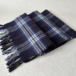 May include: A folded navy blue scarf with a plaid pattern in white and blue. The scarf has fringed edges in navy and white. The scarf is made of a soft material.