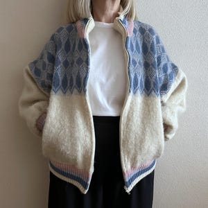 May include: A zip-up jacket with a cream-colored lower half and a blue diamond pattern on the upper half. The jacket has a cream-colored collar and cuffs with pink and blue stripes. The jacket is made of a soft, textured material.