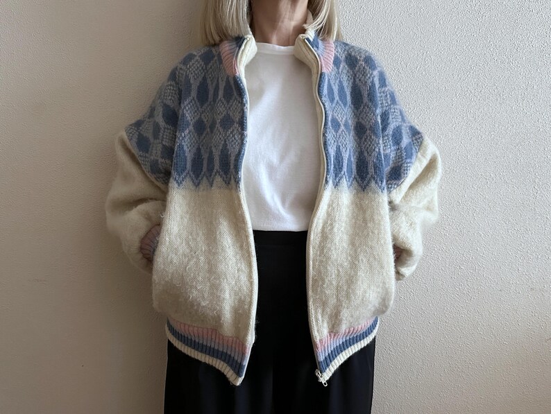 May include: A zip-up jacket with a cream-colored lower half and a blue diamond pattern on the upper half. The jacket has a cream-colored collar and cuffs with pink and blue stripes. The jacket is made of a soft, textured material.
