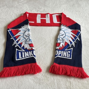 May include: A knitted scarf in red, white, and navy blue. The scarf features a lion graphic and the word "LINKÖPING" in white. The ends have red fringe, and the top section displays the letters "LHC".