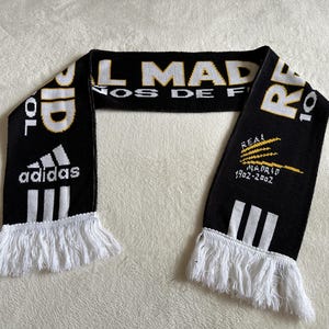 May include: Black and white Real Madrid scarf with white fringe. Features the Adidas logo, the words "REAL MADRID" in white and gold, and the years "1902-2002".