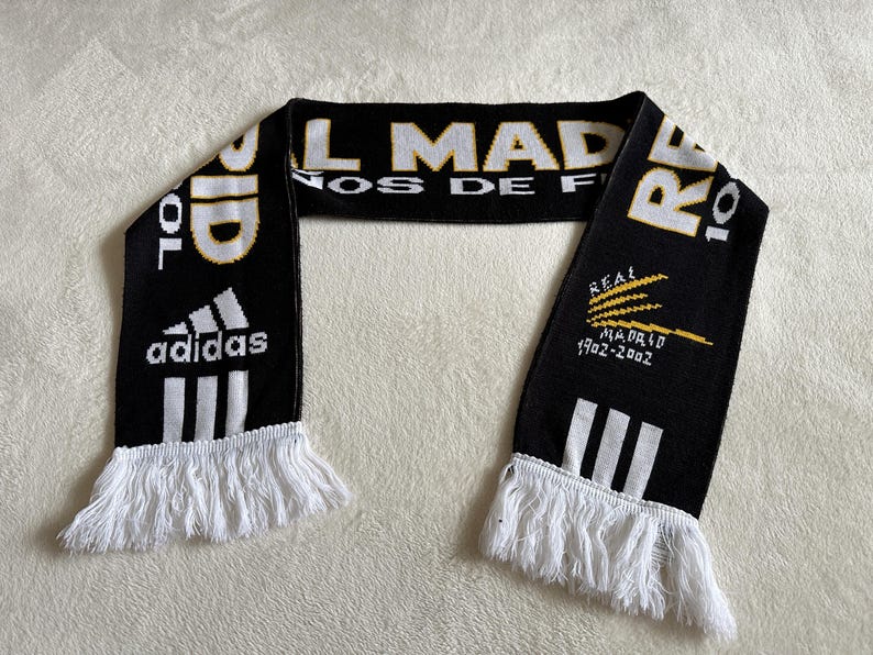 May include: Black and white Real Madrid scarf with white fringe. Features the Adidas logo, the words "REAL MADRID" in white and gold, and the years "1902-2002".