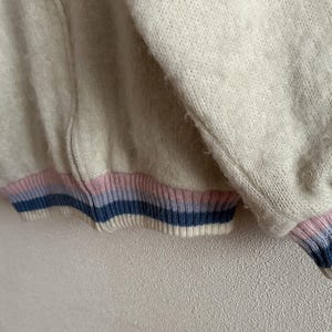 May include: Close-up of a cream-colored zip-up sweater with a ribbed cuff and hem. The cuffs and hem feature horizontal stripes in light pink, light blue, and navy blue. The sweater has a soft, textured appearance.