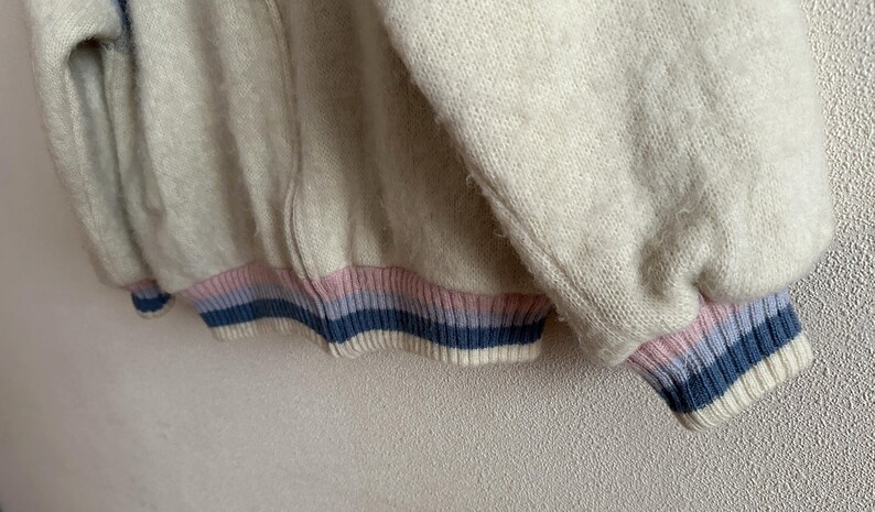 May include: Close-up of a cream-colored zip-up sweater with a ribbed cuff and hem. The cuffs and hem feature horizontal stripes in light pink, light blue, and navy blue. The sweater has a soft, textured appearance.