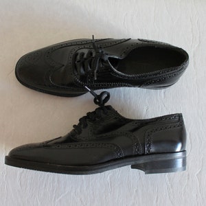 May include: A pair of black leather oxford shoes with brogue detailing. The shoes have a lace-up closure and a rounded toe.