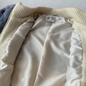 May include: Cream-colored zip-up jacket with a blue and white plaid pattern on the sleeves. The jacket has a cream-colored ribbed collar and cuffs. The interior is lined with a smooth, off-white satin fabric. A tag is visible inside.
