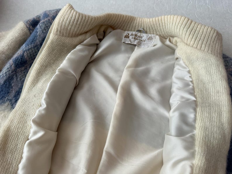 May include: Cream-colored zip-up jacket with a blue and white plaid pattern on the sleeves. The jacket has a cream-colored ribbed collar and cuffs. The interior is lined with a smooth, off-white satin fabric. A tag is visible inside.