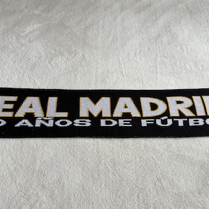 May include: Black scarf with white and gold text that reads "REAL MADRID" and "100 A&Ntilde;OS DE F&Uacute;TBOL". The scarf is likely made of a soft material and is displayed on a white, textured surface. The scarf is a sports fan item.