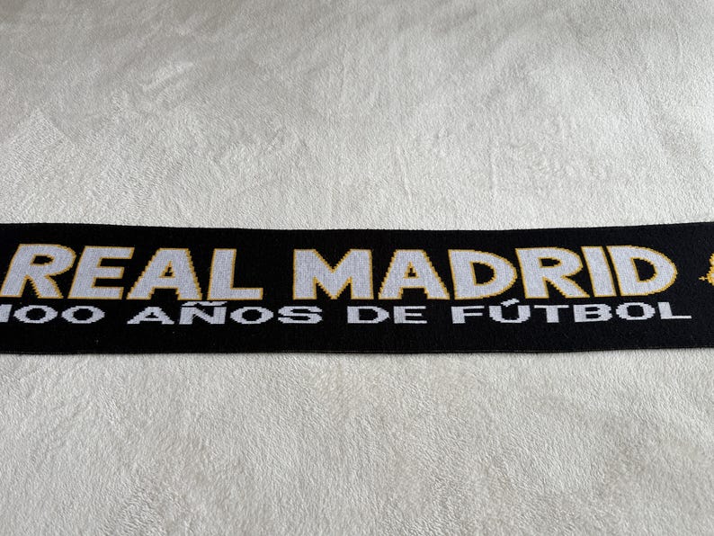 May include: Black scarf with white and gold text that reads "REAL MADRID" and "100 A&Ntilde;OS DE F&Uacute;TBOL". The scarf is likely made of a soft material and is displayed on a white, textured surface. The scarf is a sports fan item.