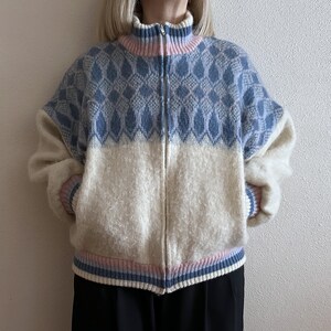 May include: A zip-up sweater with a high collar. The top half is light blue with a diamond pattern, and the bottom half is off-white. The collar and cuffs have pink and blue stripes. The sweater is made of a soft, textured material.