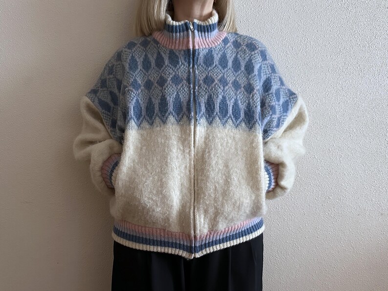 May include: A zip-up sweater with a high collar. The top half is light blue with a diamond pattern, and the bottom half is off-white. The collar and cuffs have pink and blue stripes. The sweater is made of a soft, textured material.