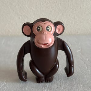 Puede incluir: Un mono de juguete de plástico marrón con la cara rosa y ojos grandes. El mono tiene brazos largos y está de pie sobre una superficie blanca.
