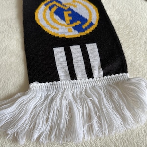 May include: Black and white knitted scarf with a team logo and three white stripes. The scarf has white fringe at the bottom. The team logo is a circular emblem with a blue and white design.