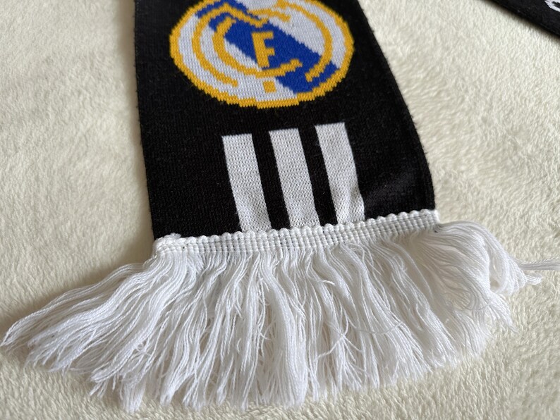 May include: Black and white knitted scarf with a team logo and three white stripes. The scarf has white fringe at the bottom. The team logo is a circular emblem with a blue and white design.